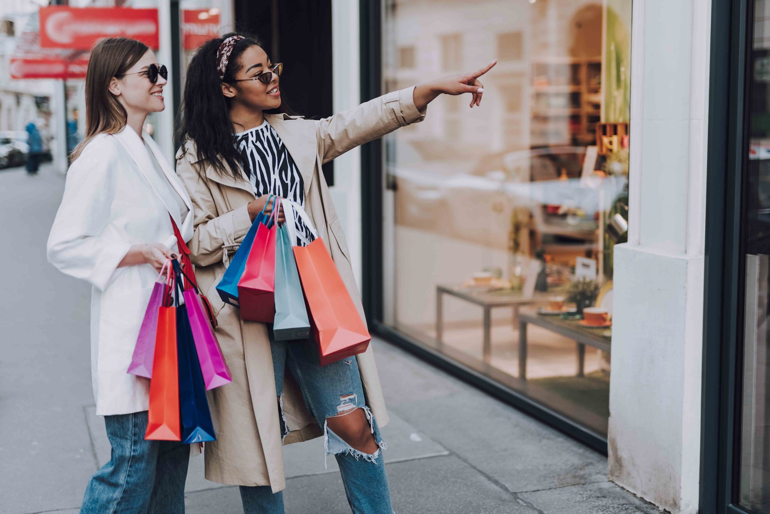 two woman holding shopping bags looking and pointing at a store front