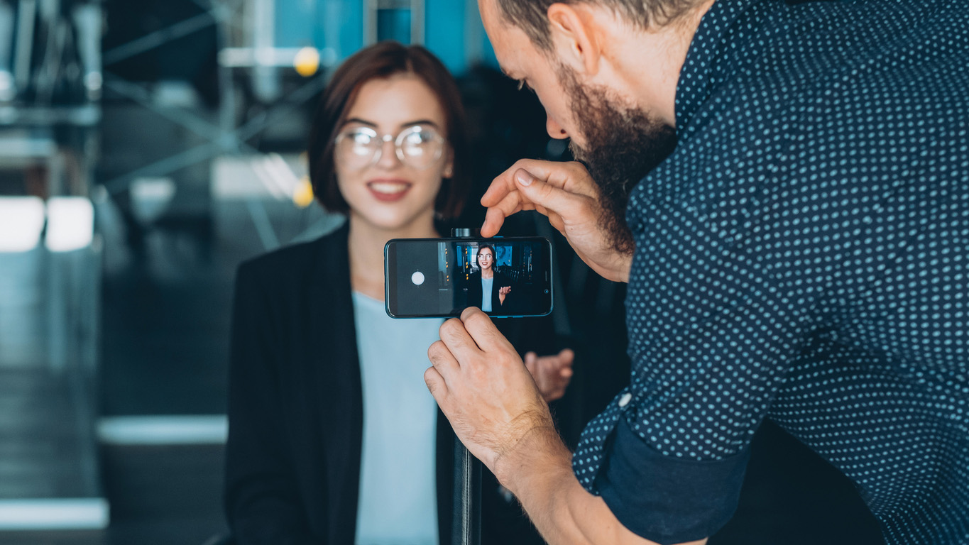 A woman in business casual attire sits in an office, as a man sets up on a phone on a tripod in front of her to film a video for social media.